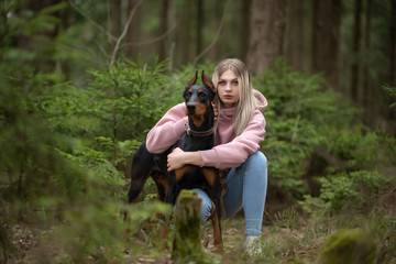 Fair-haired girl hugs a Doberman in the forest. Close-up photo.