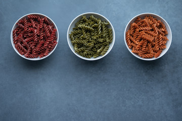 Three white bowls with different types of uncooked vegetable pasta made of spinach, broccoli, lentil, red bell pepper, tomato, pumpkin, butternut squash. Top View.