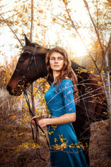 Naklejka premium Beautiful girl with her wavy hair in a blue dress looks into the distance. A bay horse stands behind in a green meadow in the sunset. Magical, fabulous fantasy photography.