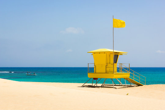 Colorful lifeguard station off duty on empty beach in Corralejo natural park, Fuerteventura. Small fishing boat on turquoise sea with yellow flag on top of baywatch tower on sunny day