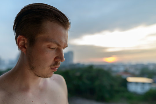 Face of young shirtless man thinking and looking down against view of the city