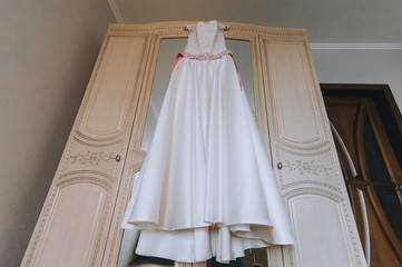 Obraz na płótnie Canvas Wedding white long dress of the bride hangs on a hanger, on a wooden wardrobe with a mirror in the hotel room. Photography, concept.