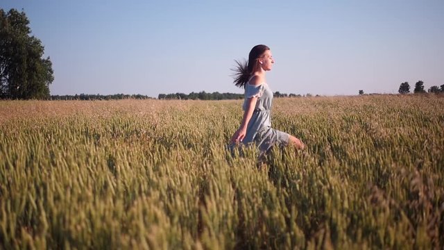 Happy young girl runs in slow motion across the field, touching her ears of wheat. Beautiful free woman enjoying nature in warm light in a wheat field at sunset background. Girl travels.
