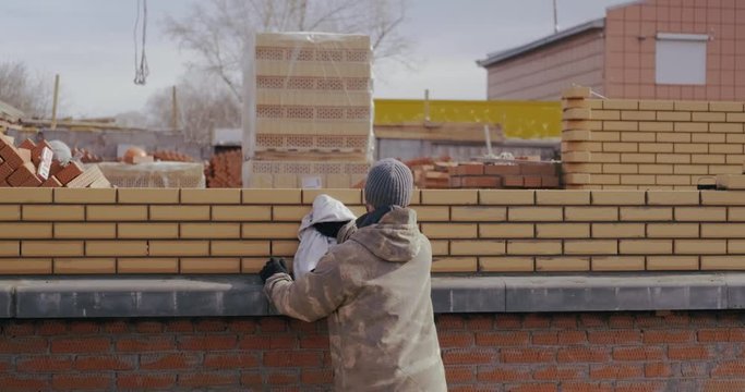 Professional builder in khaki uniform wipes brick wall after his work. Worker overwrites brick wall. Builder wipes excess solution between seams. Builder is building house. Construction of building.