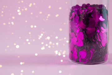 Glass bottle with purple heart shaped sparkles on pink background with copy space and blurred lights bokeh, defocused. Festive concept for your creative design