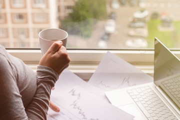 Woman freelancer working on laptop with graphics and charts printed on the paper standing near the window of her apartment. Girl drinking coffee early in the morning. remote work