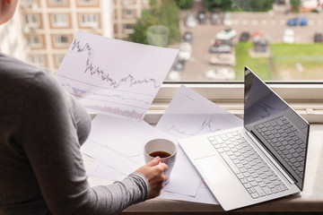Woman freelancer working on laptop with graphics and charts printed on the paper standing near the window of her apartment. Girl drinking coffee early in the morning. remote work
