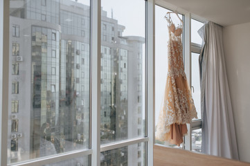 A long cream wedding dress is hanging on a hanger near a window in a beautiful interior, against the backdrop of city buildings. Morning and preparation of the bride. Photography, concept.