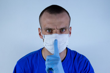 Young man in a protective mask on a blue background. Warns, Holds index finger in glove at mouth.