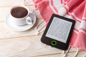 Cup of hot coffee near e-book reader over red cotton stole on a white wooden surface. E-reading for pleasure and education.