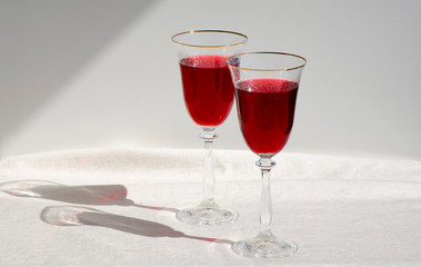 Two crystal glasses with red wine. Glass goblets with alcohol against background of white wall. White linen tablecloth. Direct sunlight, long shadows. Selective focus image. Copy space.
