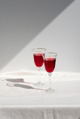 Two crystal glasses with red wine. Glass goblets with alcohol against background of white wall. White linen tablecloth. Direct sunlight, long shadows. Selective focus image. Copy space.