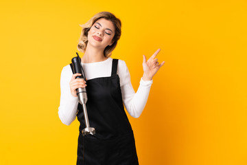 Girl using hand blender isolated on yellow background pointing finger to the laterals and happy