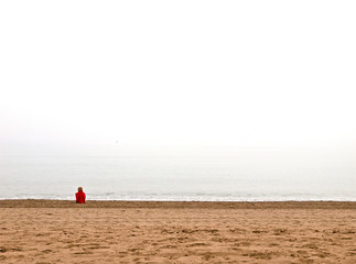 Una mujer con jersey rojo mira en horizonte del mar sentada en la arena meditando sobre el futuro