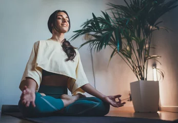 Foto op Canvas Young happy beautiful woman in cozy cropped sweatshirt and leggings practicing yoga at home sitting in lotus pose on yoga mat meditating smiling relaxed with closed eyes Mindfulness meditation concept © iana_kolesnikova