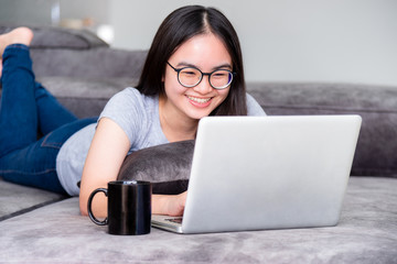 Portrait beautiful Asian young woman lying relax work with a laptop, Cute girl teens looking at the monitor smile happily on the sofa working remotely with an internet computer communication from home