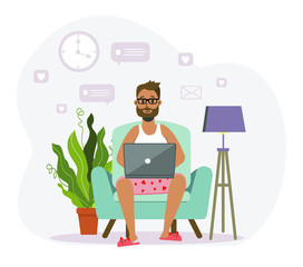 A bearded man sits on a sofa in family shorts and slippers, and works at a computer, laptop