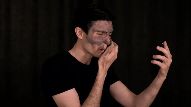 Caucasian man in black clothes applies cosmetic clay on his face while explaining to the phone screen, on a black background, portrait.