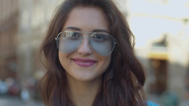 Close up of pretty young Caucasian brunette woman with long dark hair and in sunglasses smiling cheerfully to camera outdoor. Portrait of beautiful happy girl standing at street and laughing joyfully.