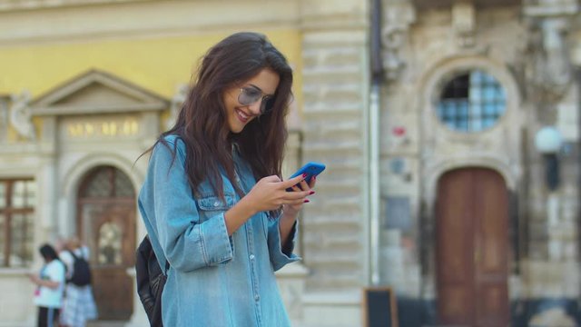 Joyful pretty young Caucasian girl in sunglsses laughing and chatting while texting message on smartphone in city center. Attractive brunette woman tapping, typing and scrolling on mobile phone.