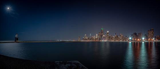 A beautiful panoramic shot of the Chicago skyline and city at night from the curved pier at North Avenue beach with light radiating from the moon and lights reflecting on the water of Lake Michigan.