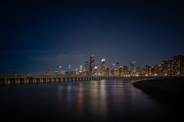 Obraz na płótnie Canvas Colorful building lights reflecting off of standing water adjacent to corrugated steel sheet piling and concrete pier with the Chicago Skyline on the horizon and stars in the dark sky above.