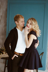 The guy and the girl are having supper at festive table. table is decorated in New Year's style. Blonde in black dress, with red lipstick on her lips. Cutlery. Blue wall. Decorated Christmas tree.