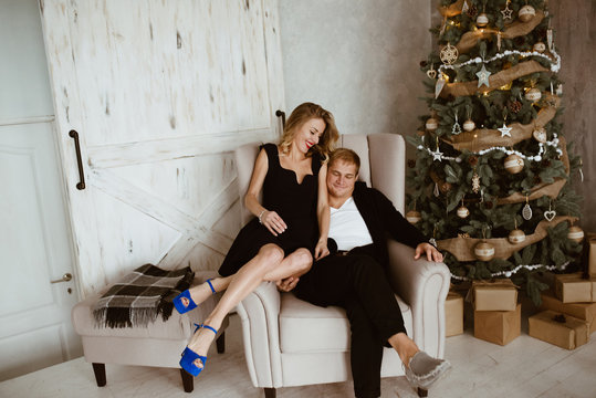 A guy and a girl near the New Year tree. Blonde in a black evening dress and with red lipstick on her lips. Beige chair. Blue sandals. Gifts under the Christmas tree. White wooden doors.