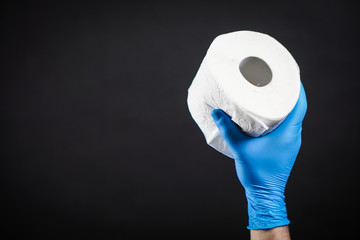 Close-up detail on a man's hand in a blue rubber glove as he holds a white roll of toilet paper on the black background. The topic is about Corona virus - COVID 19.