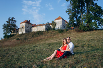 Naklejka na ściany i meble Lovely couple sitting on the grass and hugging outside. Beautiful young woman in red dress with her boyfriend
