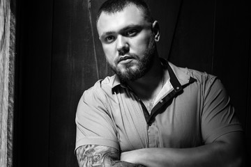 brutal bearded man with a tattoo on his arm, a portrait of a man in dramatic light against a brown wooden wall, attractive bearded male with tattoo on arm dressed in a shirt, man looks in the side