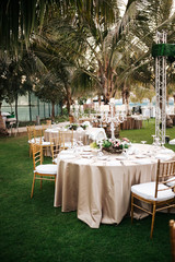 International Wedding outdoor celebration party under palm trees. Served tables on green area in hotel. Landyard. Beige and pink colors. Close-up and wide angle.