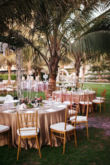 Fototapeta na wymiar International Wedding outdoor celebration party under palm trees. Served tables on green area in hotel. Landyard. Beige and pink colors. Close-up and wide angle.