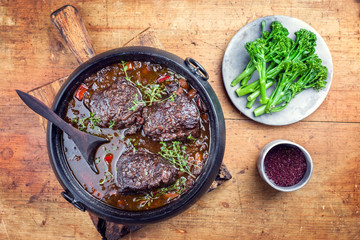 Traditional German braised beef cheeks in brown red wine sauce with carrots and broccoli offered as top view in a modern design stewpot on an old rustic board
