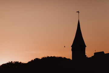 Obraz premium The silhouette of the old tower with a flag against the sunset sky background. A bird is flying in the sky.