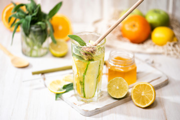 The glass of fresh lemonade with a honey. Beverage with lemon, sage, orange, and ice on the light wooden background. Summer cold drink or cocktail