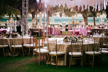 International Wedding outdoor celebration EVENING party under palm trees. Served tables on green area in hotel. Landyard. Beige and pink colors. Close-up and wide angle. - 341813390