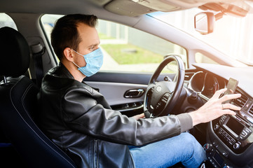 Young man in a medical mask in a car during an epidemic. He is looking for a way in the navigator on the phone on the dashboard