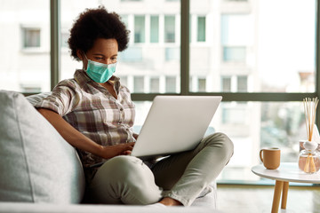 African American woman with face mask using laptop while relaxing on the sofa.