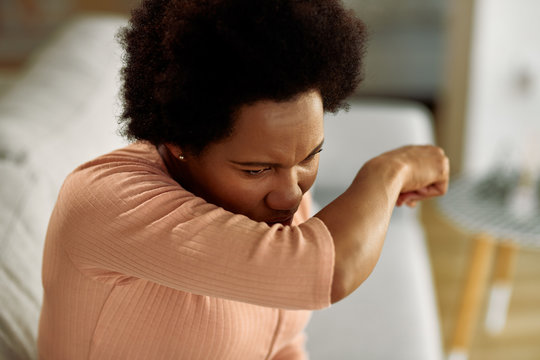Black woman with flu virus sneezing into elbow at home.
