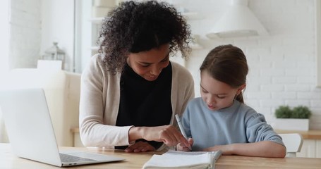 Smiling african woman tutor or foster parent mum helping cute caucasian school child girl doing homework sitting at kitchen table. Diverse nanny and kid learning writing in notebook studying at home.