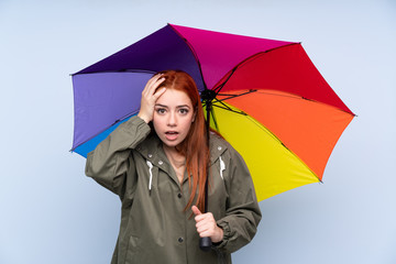 Redhead teenager girl holding an umbrella over isolated blue background with surprise facial expression