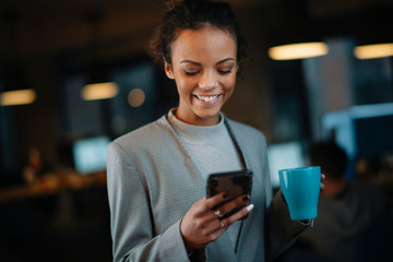 Young businesswoman using phone and drinking coffee in office.	