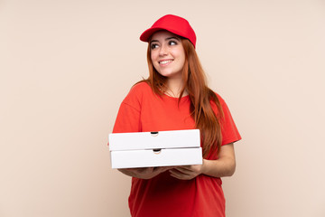 Pizza delivery teenager girl holding a pizza over isolated background laughing and looking up