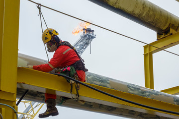 Working at height. An abseiler wearing Personal Protective Equipment (PPE) step on the structure for touch up painting activities with background flare tip burning in the sky.
