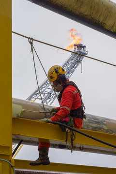 Working at height. An abseiler wearing Personal Protective Equipment (PPE) step on the structure for touch up painting activities with background flare tip burning in the sky.