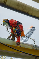 Working at height. An abseiler wearing Personal Protective Equipment (PPE) step on the pipeline to check surfaces prior to start painting activities with background flare tip burning in the sky.