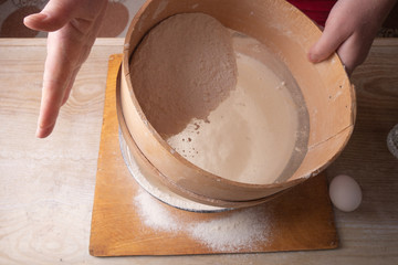 Large bowl, cutting board, sieve for sifting flour on the table. Female hands sifting flour in the home kitchen.