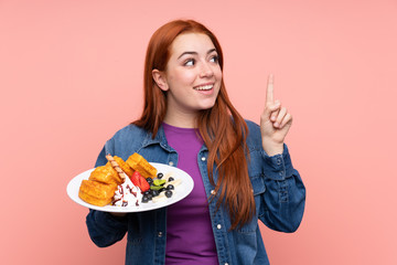 Redhead teenager girl holding waffles over isolated pink background intending to realizes the solution while lifting a finger up