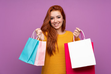 Fototapeta na wymiar Redhead teenager girl over isolated purple background holding shopping bags and smiling